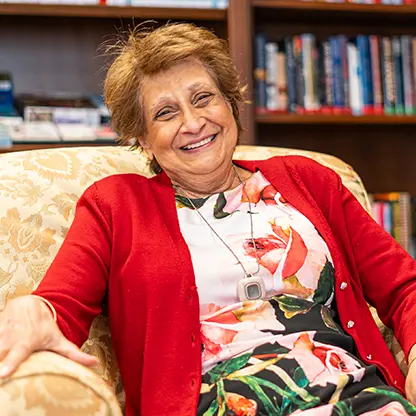 A happy, smiling senior woman wearing a floral patterned dress and red cardigan sitting in on a comfortable chair