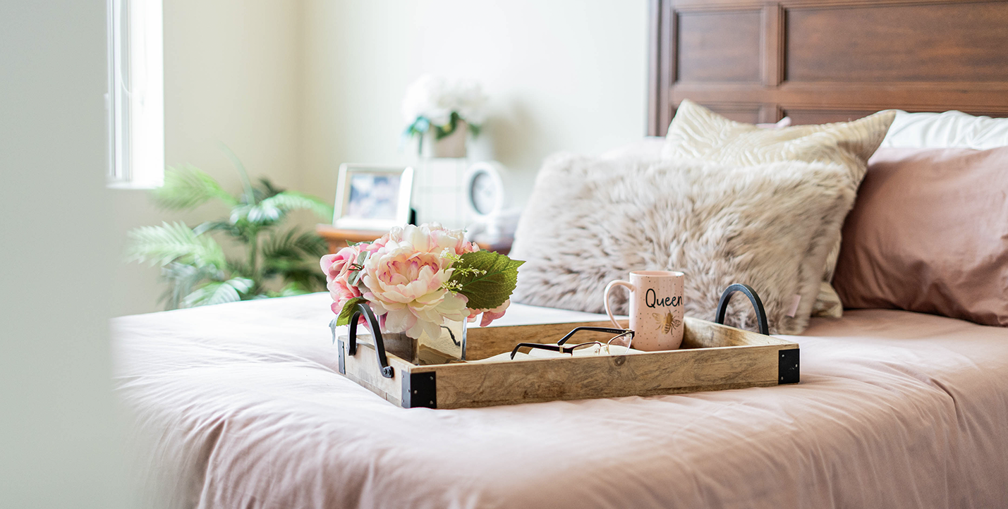 An image of a comfortable looking bed with a pink comforter set on it, and an elegant tray sitting near the edge of the bed.