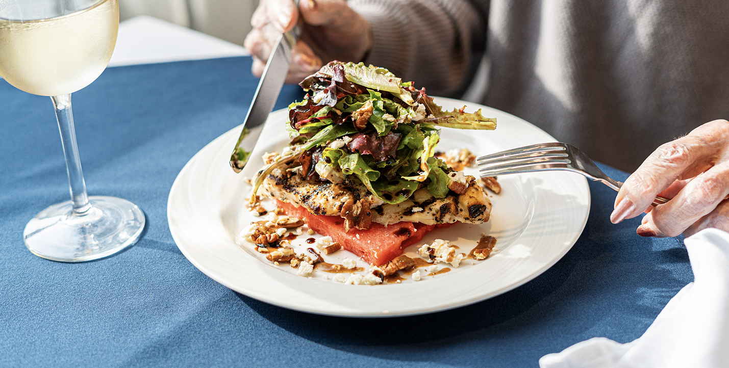 A white plate with a colourful meal on it featuring a grilled chicken breast stacked on top a rectangular slice of watermelon, greens, and nuts.
