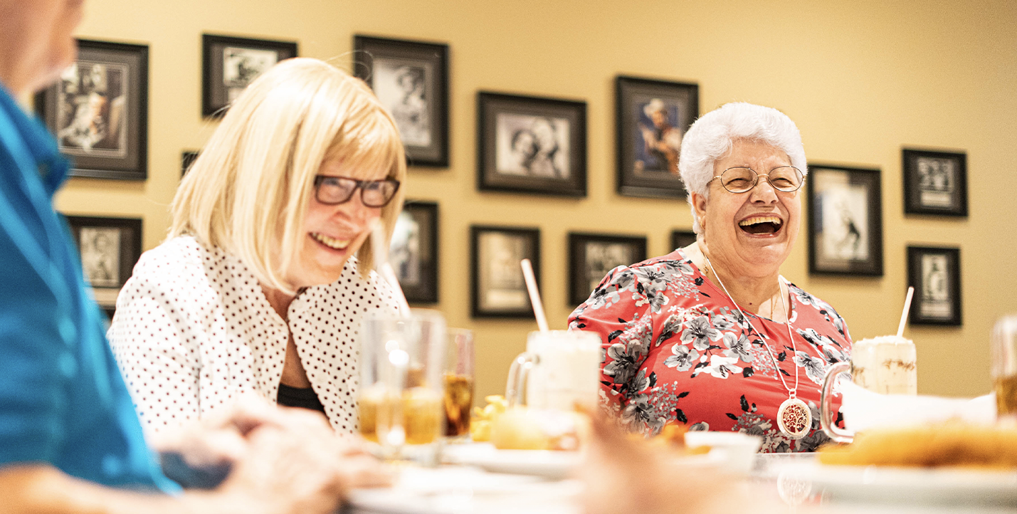 Seniors sitting at a 1950s era diner, laughing while eating fries, burgers, and drinking milkshakes