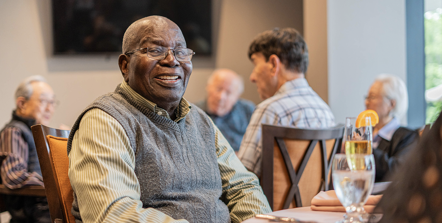 A senior black male smiling at the camera, enjoying the company of other seniors in a busy dining room