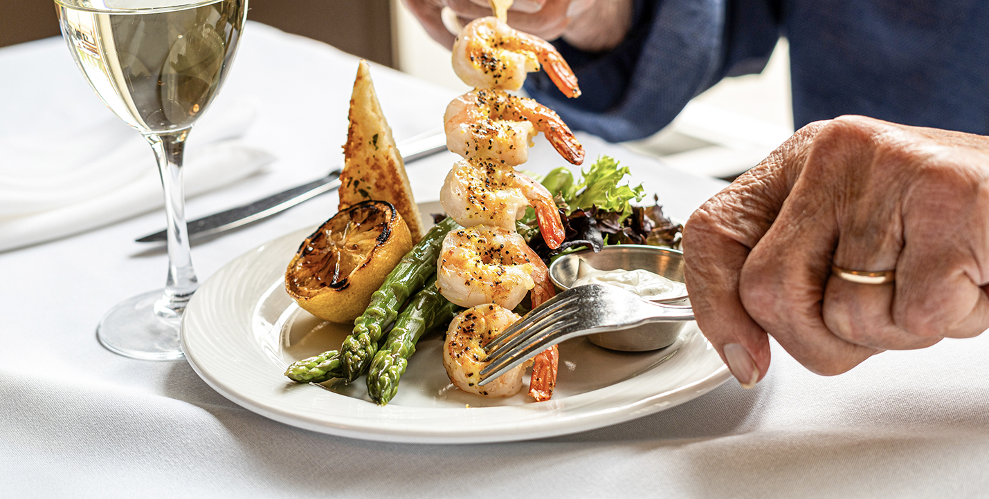 A senior using a fork to remove a shrimp from a wooden skewer. The plate features delicious grilled shrimp with a side of asparagus